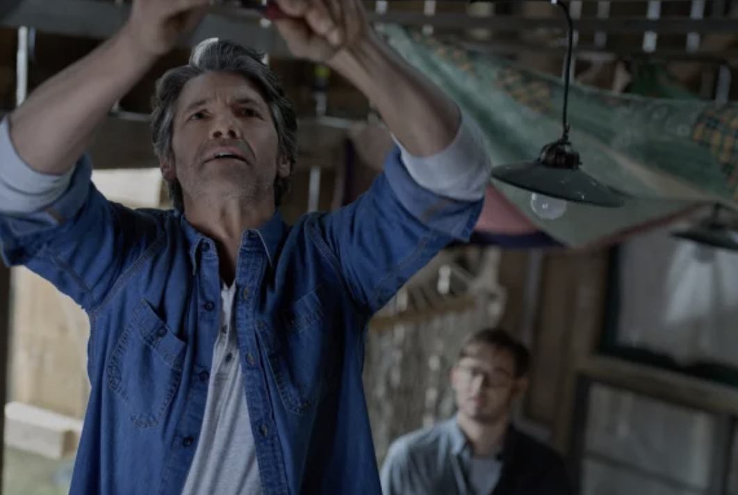 Colton Landry stands in the foreground in a barn repairing a piece of equipment. Eliot Augustine looks on in the background. 